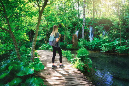 Young woman with backpack walking along a wooden path near stunning waterfalls, green trees in Plitvice Lakes national park, Croatia at sunset in summer. Female tourist. Sporty girl in forest. Hiking