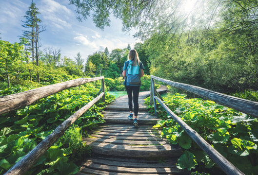 Young woman with backpack walking along a wooden boardwalk near waterfalls, green trees at sunset in summer. in Plitvice Lakes national park, Croatia. Female tourist. Girl on stairs in forest. Hiking