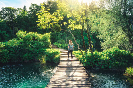 Young woman with backpack walking along a wooden boardwalk near waterfalls, green trees in Plitvice Lakes national park, Croatia at sunset in summer. Female tourist. Sporty girl in forest. Hiking
