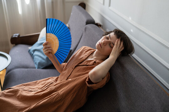 Stressed woman suffering from summer heat and migraine at home, lying on sofa waving paper fan for cooling, unwell female on couch with headache touching forehead and fanning herself with hand fan.