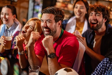 Ecstatic friends celebrating a soccer goal with a man in a red jersey at a pub. © Mediteraneo