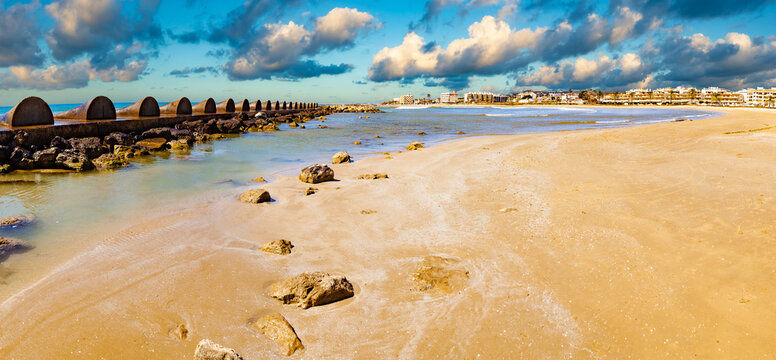Playa de arena con aguas tranquilas de color turquesa y rompeolas rocoso bajo un cielo azul nublado. Paisaje costero en Cubelles, Costa Dorada, Barcelona. Playa mediterr&aacute;nea con paisaje costero ideal 