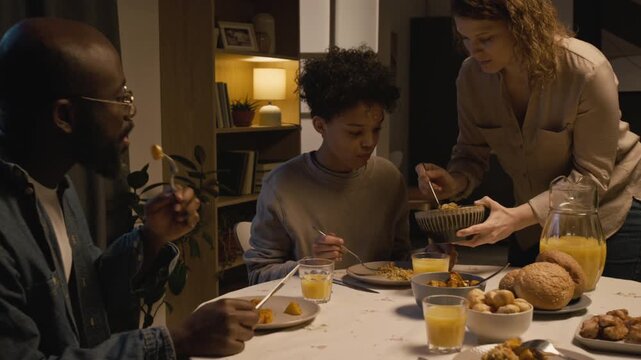 Medium shot of multiracial family sitting at dinner table together in evening and enjoying meal with mother caring for son and serving food