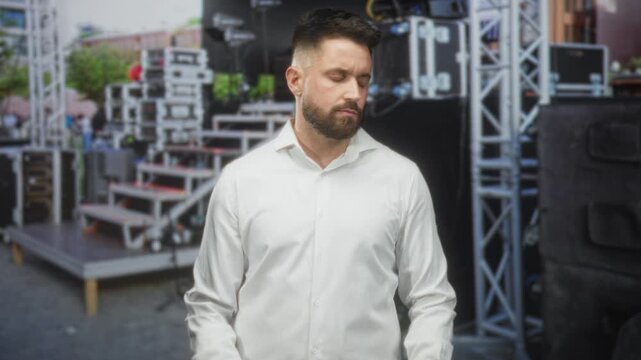 Hispanic man with beard in white shirt holds open hands palms up on a street stage near speakers and metal rigging; contemplation doubt.