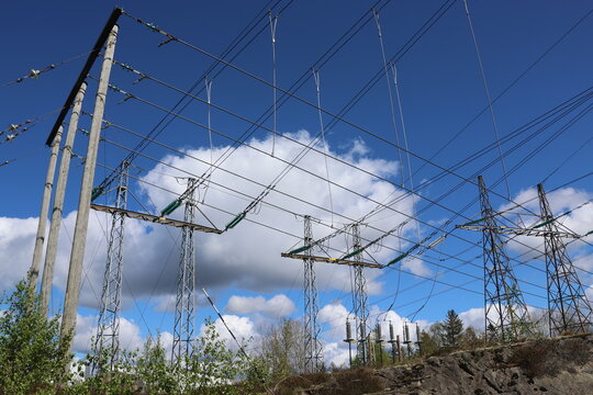 Detailed view of a high-voltage power line, captured against a clear blue sky with bright sunlight and scattered cumulus clouds. Contemporary Swedish energy infrastructure presented in sharp daylight 