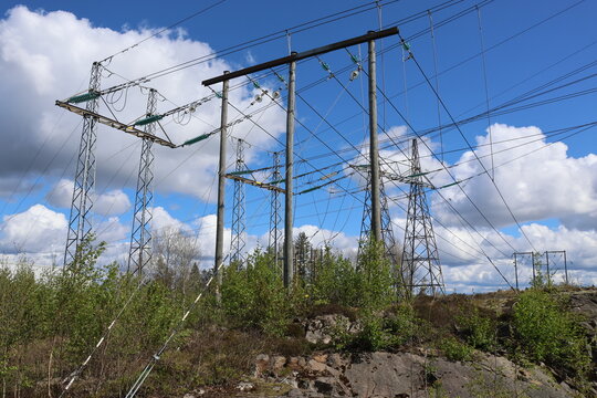 Detailed view of a high-voltage power line, captured against a clear blue sky with bright sunlight and scattered cumulus clouds. Contemporary Swedish energy infrastructure presented in sharp daylight 