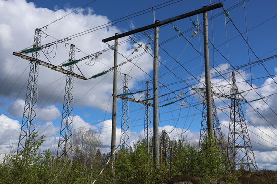 Detailed view of a high-voltage power line, captured against a clear blue sky with bright sunlight and scattered cumulus clouds. Contemporary Swedish energy infrastructure presented in sharp daylight 