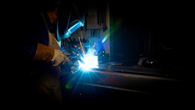 Welder in protective helmet and leather gloves welding dark steel in a dimly lit workshop. Brilliant blue-white arc flash with flying orange sparks and tool rack in background.