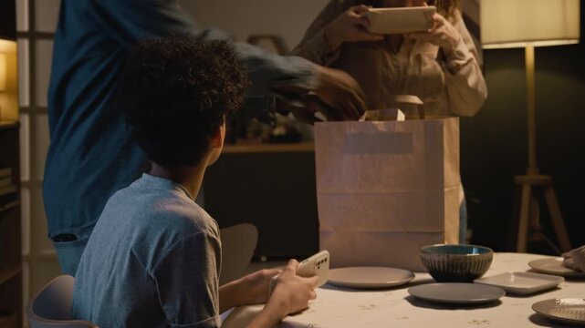 Candid scene of multiracial family setting takeout food on dinner table together with teenage boy using smartphone in foreground, lit by low lighting, copy space