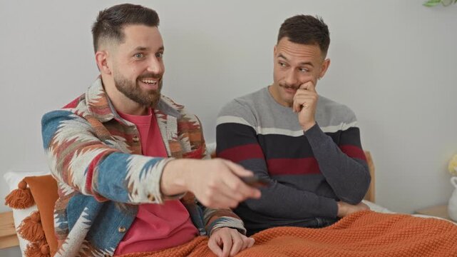 Two men sitting on a bed, one pointing a tv remote while smiling and the other rests hand on cheek in a studio; playful intimacy.