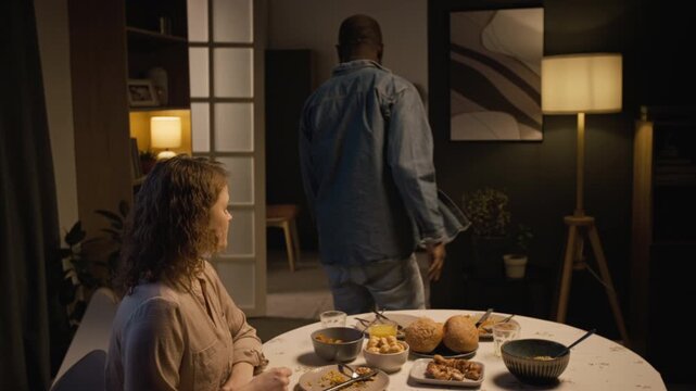 Wide angle shot of upset woman left alone at dinner table after family fight with husband leaving room angrily, scene lit by low evening light, copy space