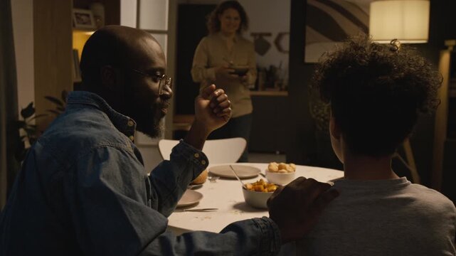 Cozy lifestyle scene of caring Black father talking to young son sitting at dinner table together as family