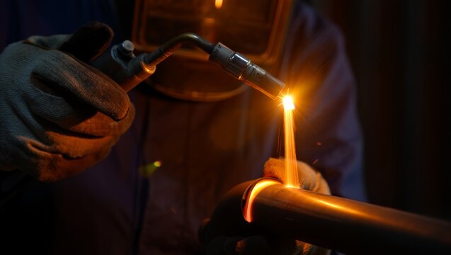 Intimate close-up of gloved hands brazing a glowing copper pipe joint with a bent MIG torch, with a bright arc flash and dripping molten orange metal captured in a dark warm hangar workshop setting.