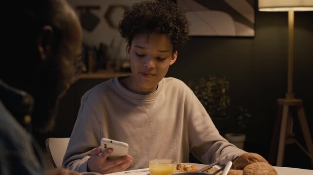 Front view shot of upset Black teenage boy checking messages online while sitting at dinner table with family, father supporting him and patting on shoulder