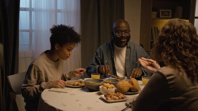 Side view shot of Black teenage boy using smartphone at dinner table with multiracial family and checking messages getting bad news, father supporting him