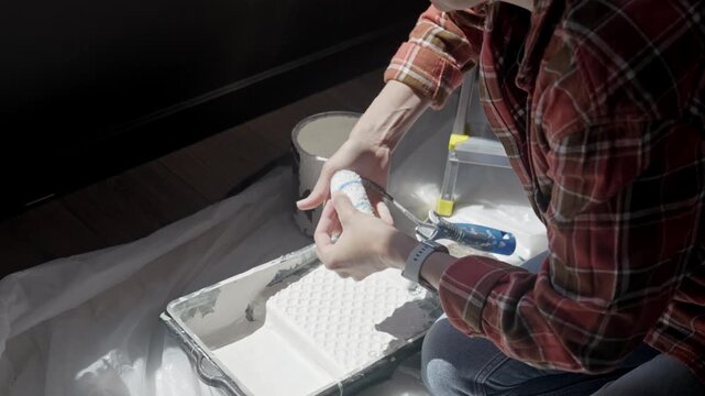 Woman fits paint roller cover onto handle near tray, can, brush and paint on protective sheet. Concept of renovation and painting preparation