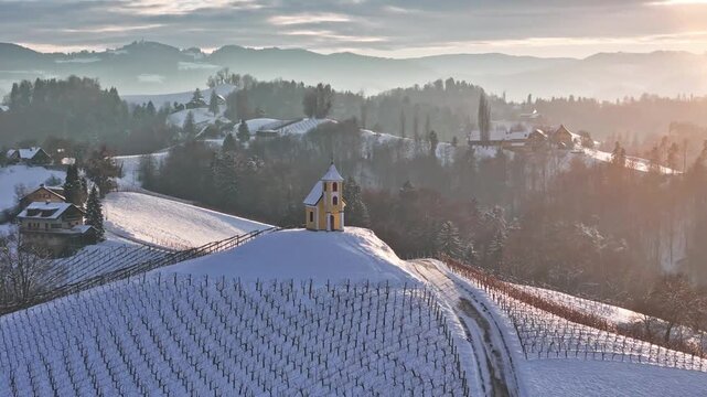 Aerial view of Dreisiebner Kapelle on a snow-covered hill surrounded by vineyards and rolling hills in Gamlitz, Styria, Austria.