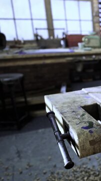 In a workshop, a wooden workbench stands with a clamp attached. Tools and materials are scattered on the table nearby. Natural light comes through large windows.