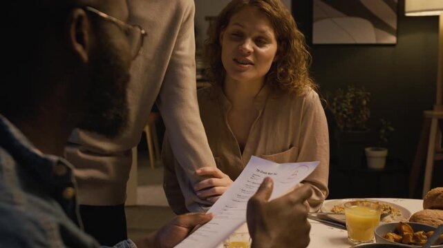 Over shoulder close up shot of Black man as father looking at sons exam papers with excellent grades at dinner table, focus flowing to smiling mother embracing son
