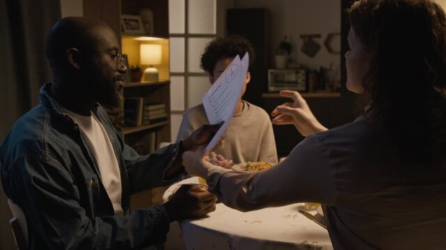 Over shoulder shot of mother showing sons exam papers to proud father during family dinner at home, scene lit with cozy evening lighting