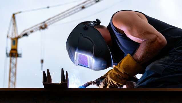 Low-angle shot of a muscular welder in a black auto-darkening helmet and leather gloves welding a steel beam on a construction site with a yellow tower crane rising against an overcast sky.