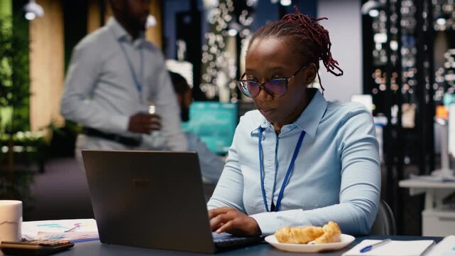 Eating disorder and emotional stress, black woman working on laptop, worker eating excessive junk food at her desk. Employee appearing worried as she handles tasks, compulsive overeating.