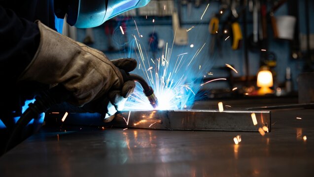 Close-up of a welder in a blue-tinted helmet and worn leather gloves MIG welding a steel box section, vivid blue arc and orange sparks scattering across a dark workshop with tool wall behind.