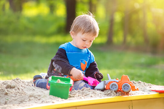 Toddler boy playing with plastic truck and shovel in sandbox at playground.