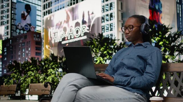 African american girl using music for work inspiration with headset and laptop on bench in the park, multitasking and ensuring productivity. Urban tech concept and peaceful life.