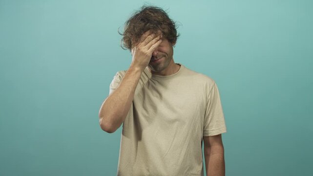 Man covering face with hand in studio wearing beige tshirt; embarrassment self reflection introspection.