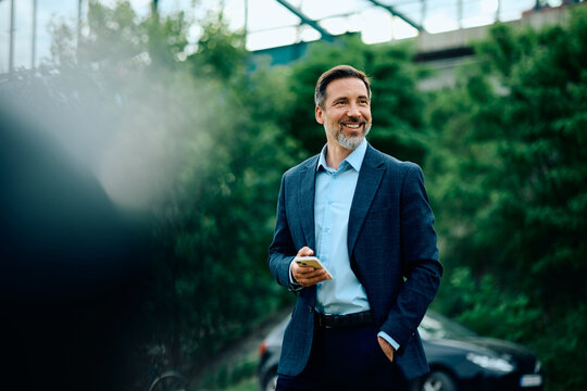 Happy entrepreneur with mobile phone outdoors looking away.