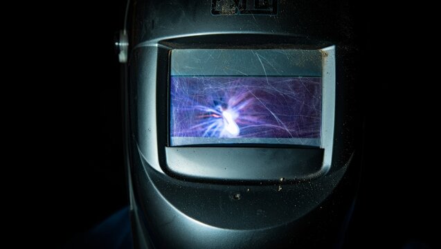 Close-up of a well-used welding helmet lens reflecting a vivid purple-blue welding arc. The scratched protective glass captures swirling plasma light against the pure black studio background.