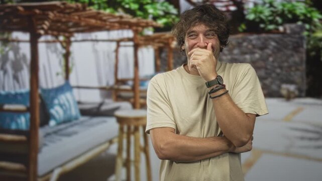 Hispanic man with hand covering mouth and crossed arms in building cabana lounge wearing a watch and bracelets near daybed and pillows; amused vacation moment.