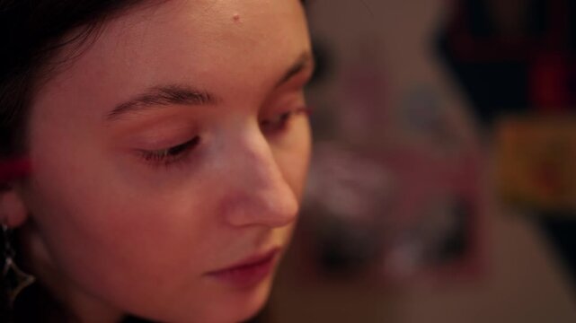 Closeup of focused woman applying red mascara with brush carefully. Precise makeup application by caucasian woman under warm light. Detailed image of female face and eyelashes. Beauty and fashion