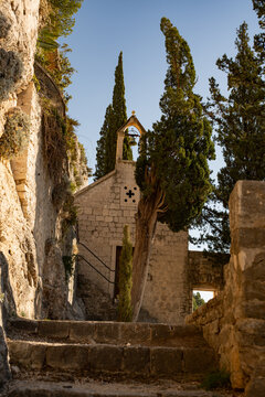 Epic Saint Jerome cave church ancient stone architecture vertical panorama. 
Cinematic low angle view of historic medieval limestone chapel facade, 
bell tower grotto, wild rocks, cypress trees and st