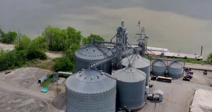 Aerial view of large metal grain silos and industrial elevators with an American flag flying near the riverbank in Aurora, Indiana, United States.