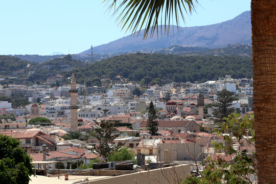 Scenic panoramic view of Rethymno old town with traditional rooftops, church minarets, and green hills under a mountain landscape, Crete, Greece.