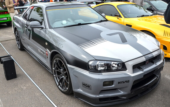 Grey Nissan Skyline GT-R R34 at an outdoor event. This JDM legend features a carbon fiber front lip and occupies a central place in automotive tuning culture