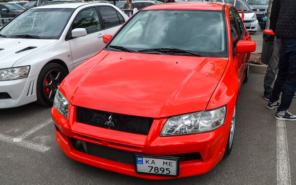Front view of a bright red Mitsubishi Lancer Evolution VII at an outdoor car meet. The JDM icon features its signature aggressive fascia and exposed intercooler