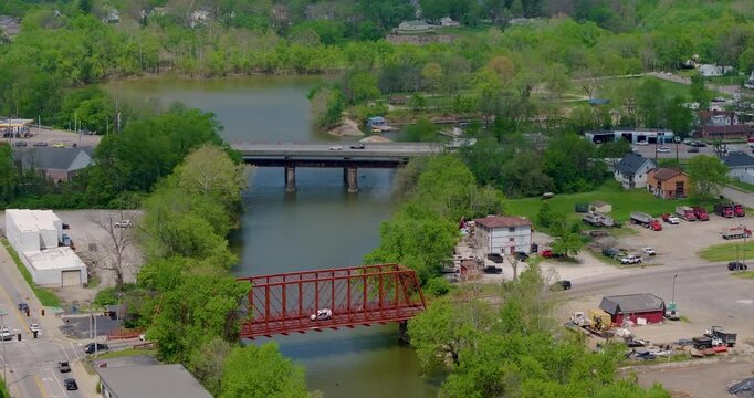 Aerial view of a red truss bridge and a modern road bridge crossing a river surrounded by lush green trees and small town buildings in Aurora, Indiana, United States.