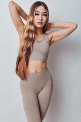 Fototapeta premium Young woman with long hair in sportswear posing with arms raised against white background for fitness and health concept. Slim body, casual style and confident expression indoors.