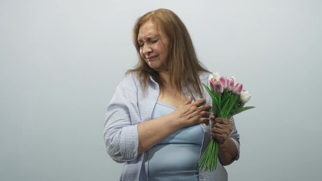 Senior woman holding pink tulip bouquet, hand on chest and eyes closed with tearful expression, wearing blue top and open shirt in studio; remembrance grief.