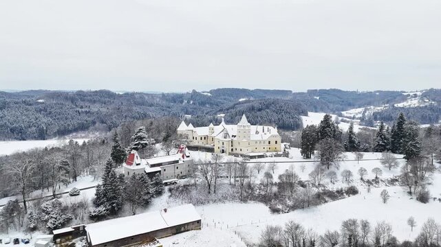 Aerial view of Schloss Kornberg with snow-covered roofs and surrounding winter forest under an overcast sky in Sudoststeiermark District, Styria, Austria.