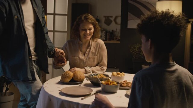 Cozy lifestyle scene of multiracial family enjoying dinner together at home with father bringing food to table and sitting down saying grace