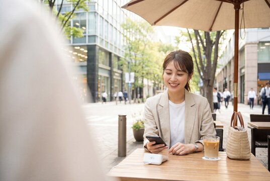 Smiling young Asian woman using a smartphone for contactless mobile payment at an outdoor cafe terrace in a modern city