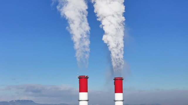 Aerial view of two red and white industrial chimneys emitting thick white smoke into a clear blue sky with distant hills in Wildon, Styria, Austria.