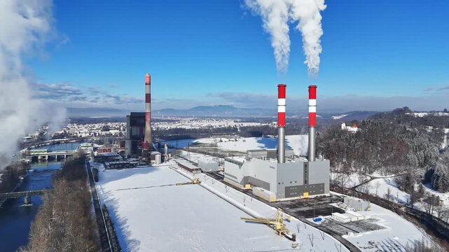 Aerial view of a power plant with tall red and white chimneys emitting smoke over a snow-covered landscape and river in Wildon, Styria, Austria.