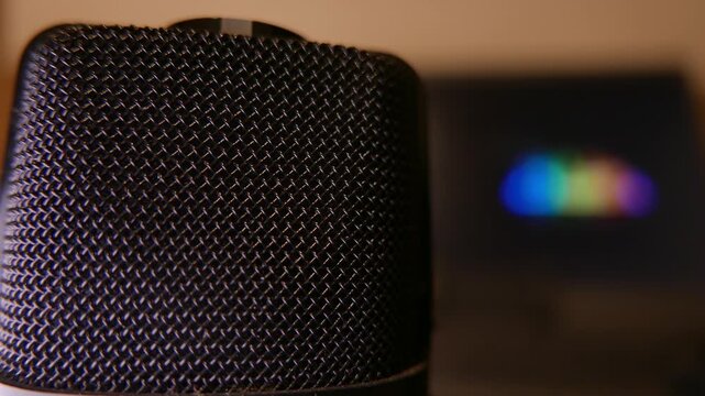 POV: recording audio. A very immersive close-up of a professional podcast microphone. In the blurred background, a laptop displays vibrant, colorful moving audio waveforms. A recording studio
