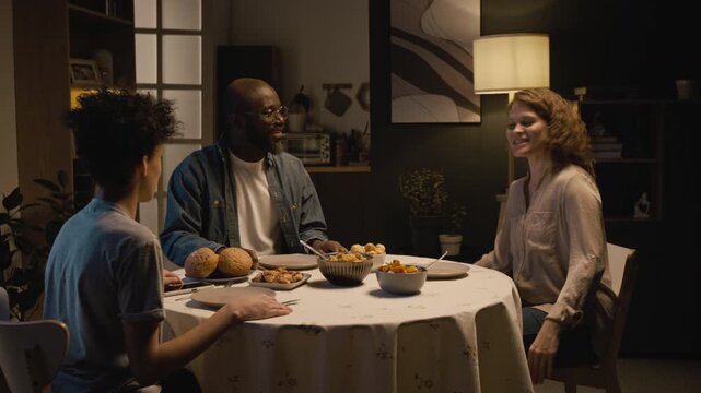 Medium shot of multiracial family enjoying dinner together at family and praying at dinner table holding hands, cozy evening scene lit by low lighting, copy space
