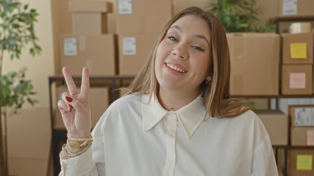 Woman showing peace sign with left hand and bracelets beside stacked cardboard packages in building; cheerful small business.